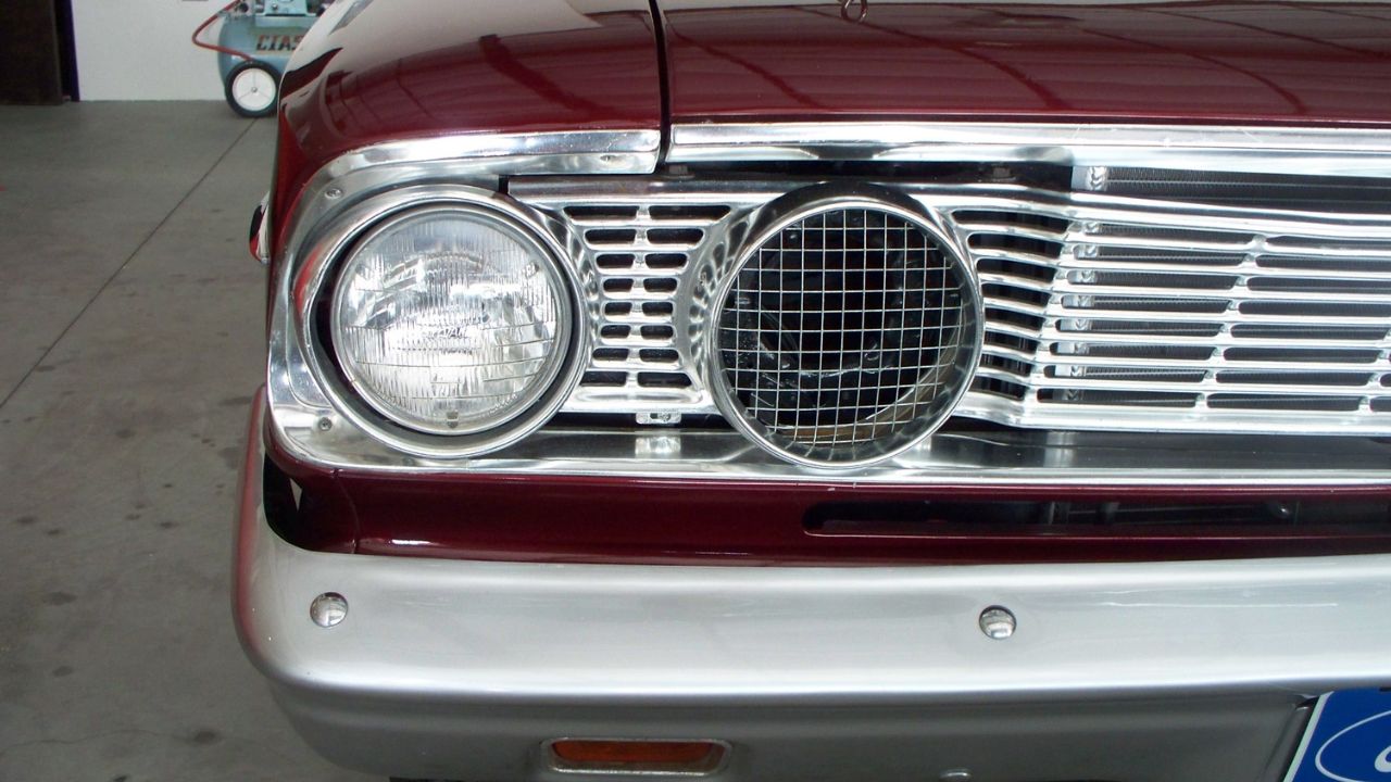 Detail of the right side engine air intake system and painted fiberglass bumper of a 1964 Ford Fairlane Thunderbolt factory experimental drag race car. The mesh-covered opening takes the place of the high-beam headlight found on a street-driven vehicle.

