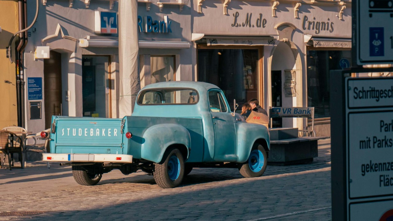 A classic Studebaker pickup truck is parked on a sunlit cobblestone street outside a bank in Europe.
