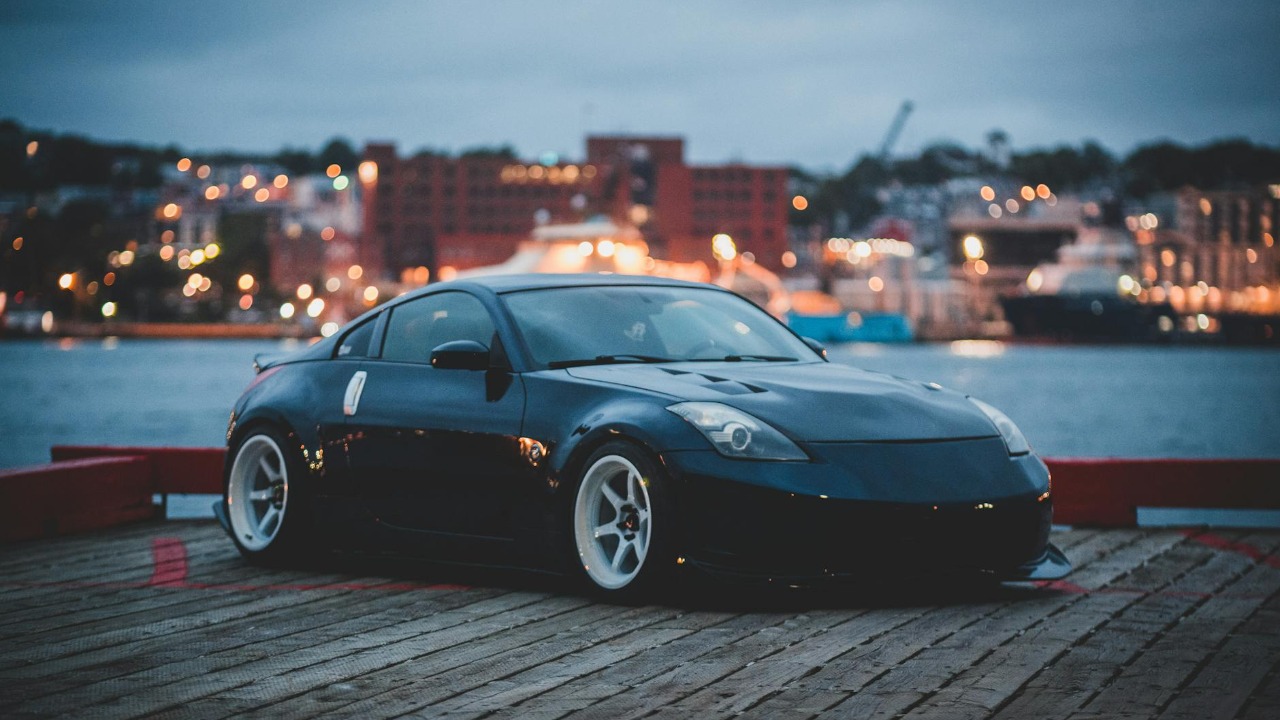 A sleek black Nissan 350Z sports car parked by the waterfront with city lights in the background at dusk.