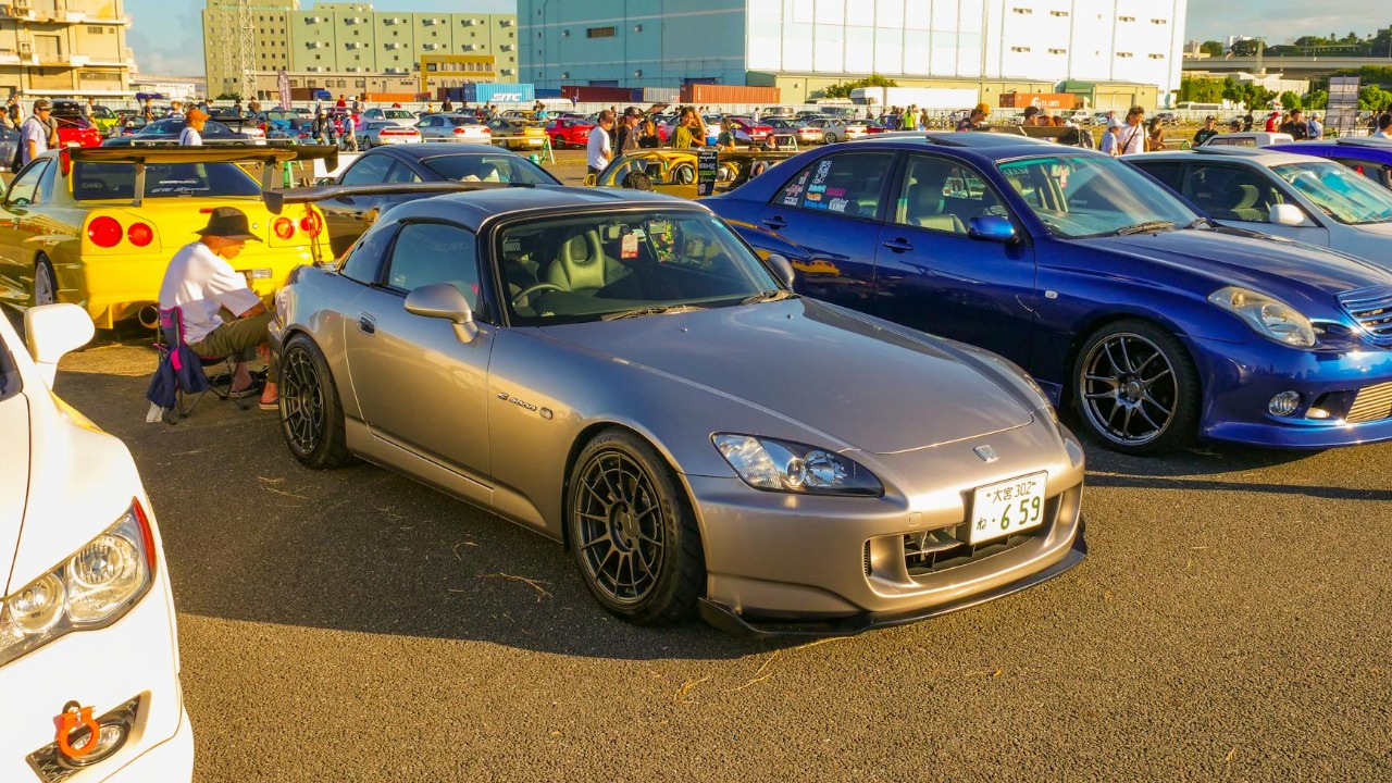 A lively car meet with JDM models including a silver Honda S2000 and other sports cars under the sunny sky.