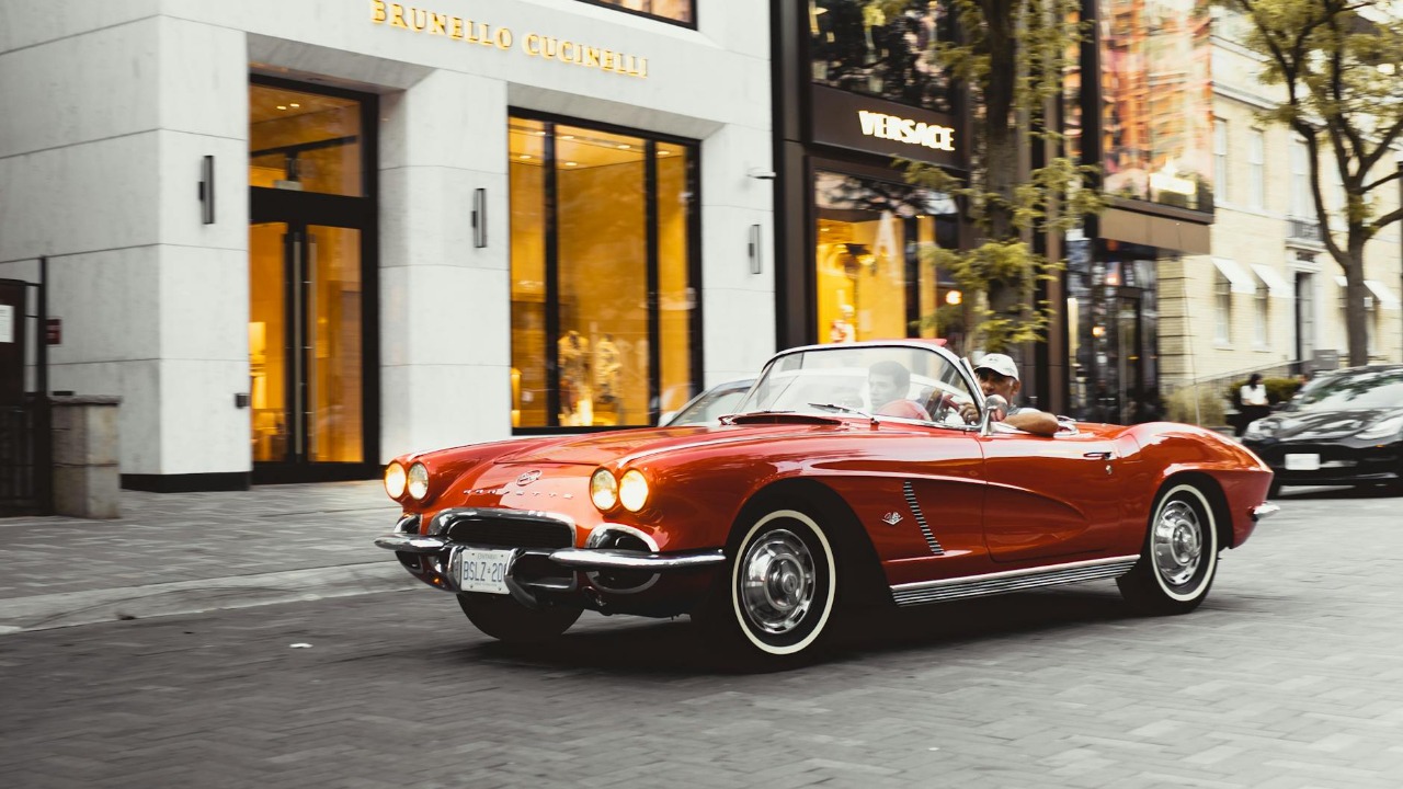Vintage red Chevrolet Corvette convertible driving past luxury shops in Toronto.