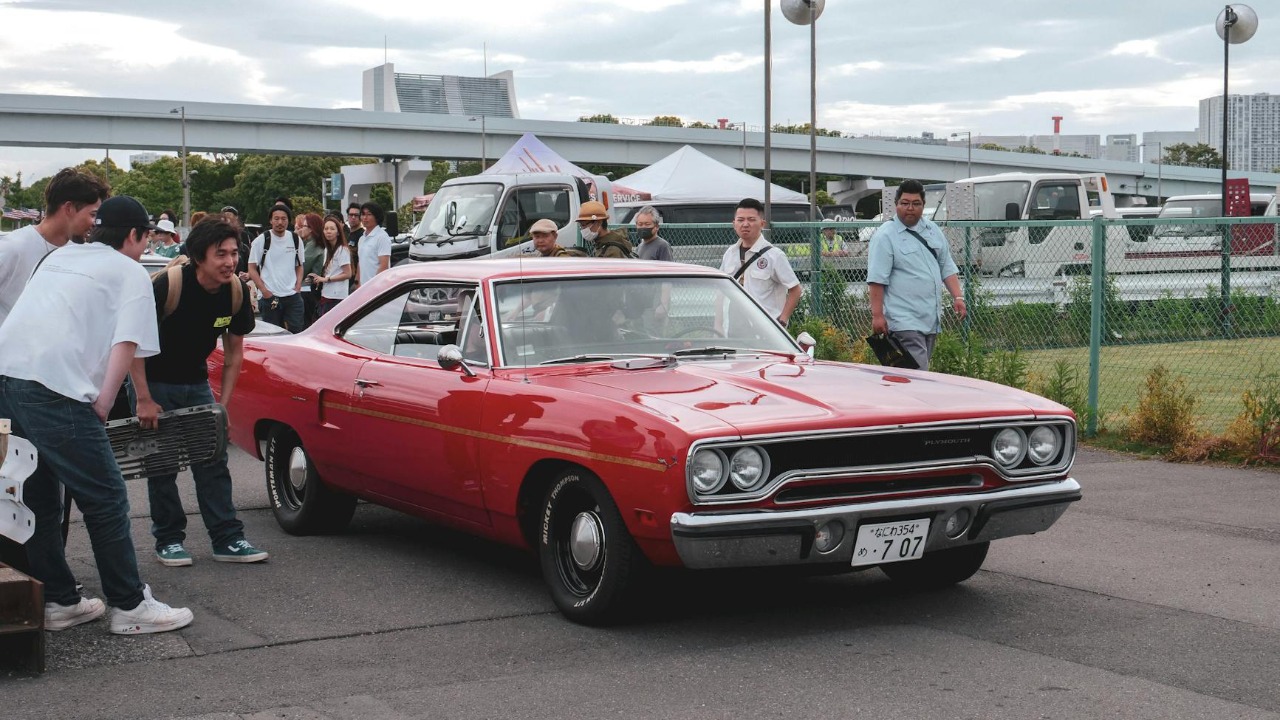 Red classic car showcased at an outdoor meetup with onlookers in summer.