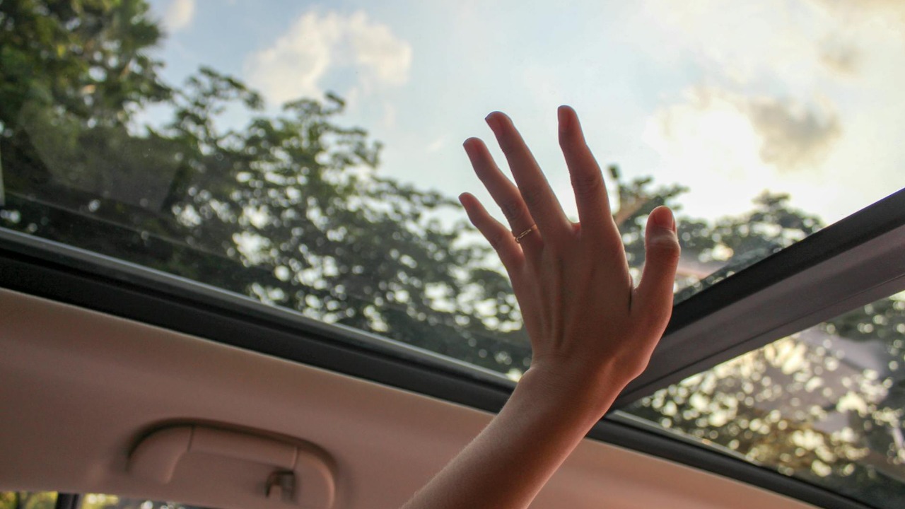 A hand reaching out through a car sunroof, touching the sky on a sunny day.