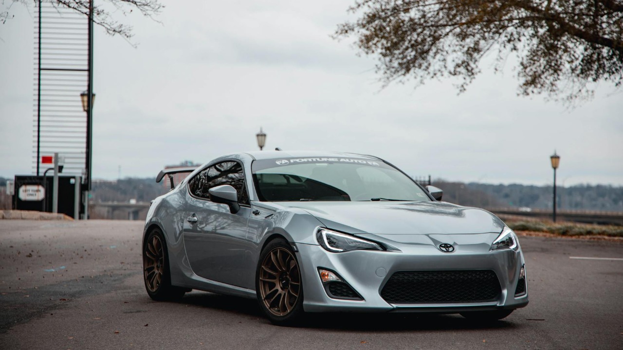 A silver sports car parked on an asphalt road surrounded by sparse trees under a cloudy sky.