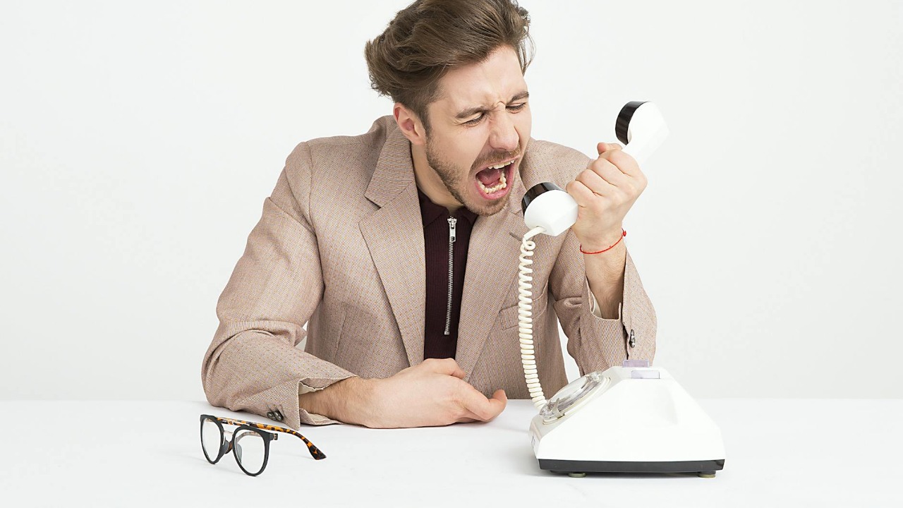 Businessman in a beige suit shouting into a vintage telephone, expressing frustration at work.