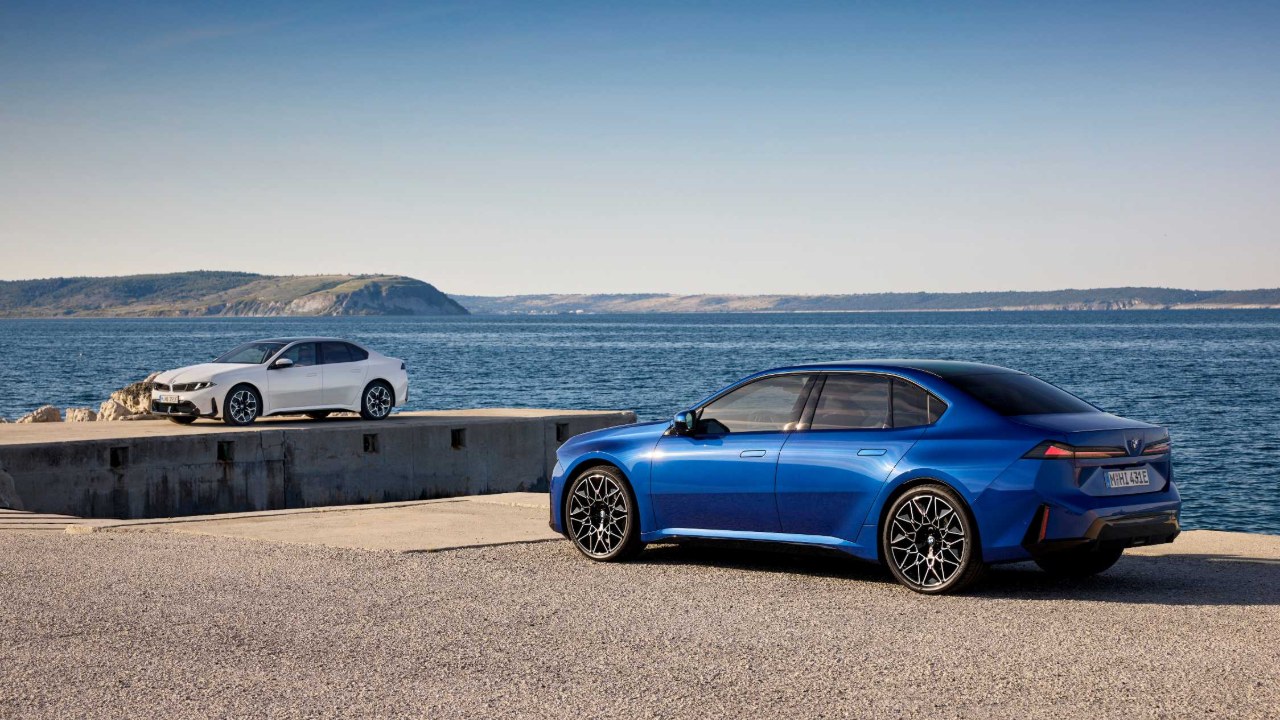 A blue 2027 BMW 3 Series sedan parked by the sea, rear three-quarter view